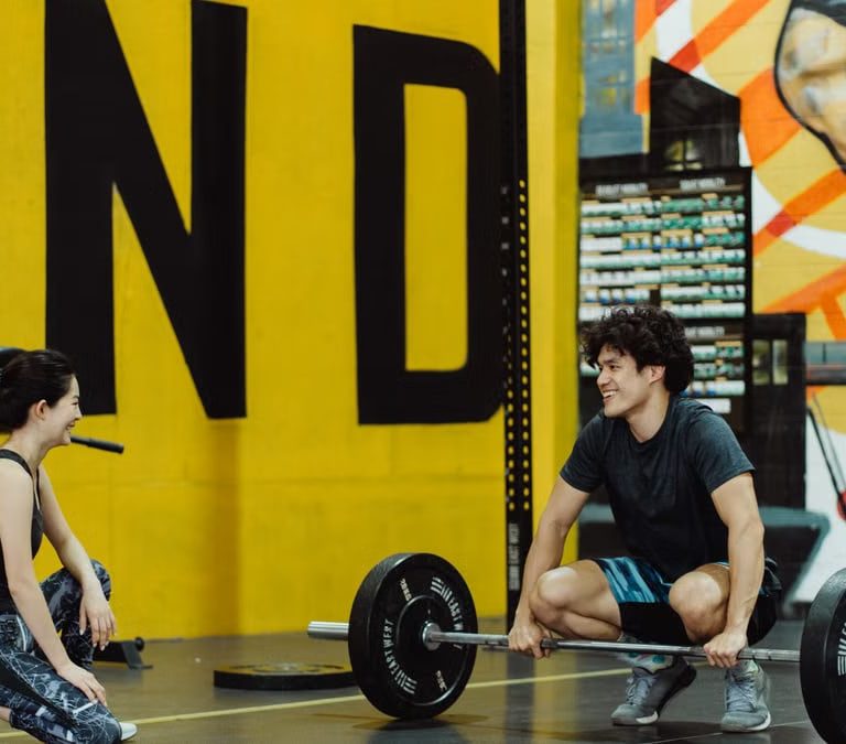 Two young adults in a gym participating in a weightlifting session, sharing a positive moment.